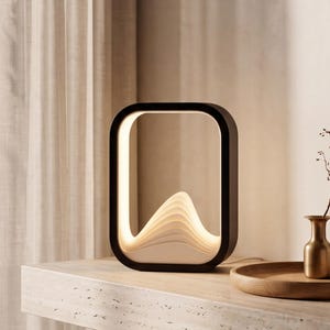 Puede incluir: Una lámpara de mesa rectangular con un marco exterior negro y una estructura interior blanca en capas, similar a ondas, que emite un brillo suave y cálido. La lámpara presenta un diseño liso con esquinas redondeadas.
