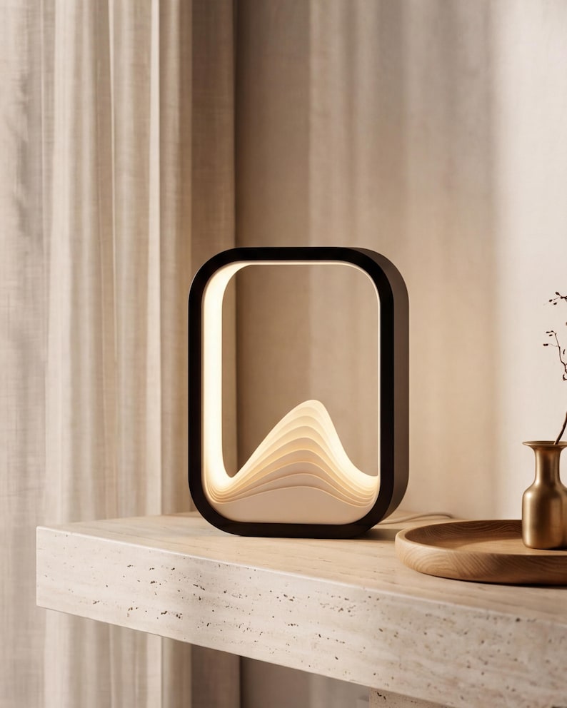 May include: A rectangular table lamp with a black outer frame and a white, layered, wave-like interior structure that emits a soft, warm glow. The lamp features a smooth, rounded-corner design and sits upright on a flat surface.