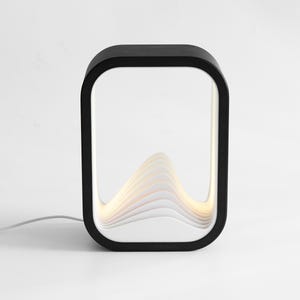 May include: A rectangular table lamp with a black outer frame and a white interior featuring layered, wave-like panels that create a mountain-shaped silhouette. The lamp emits a soft, warm glow from within the structure and includes a white power cord.