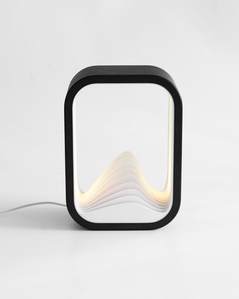 May include: A rectangular table lamp with a black outer frame and a white interior featuring layered, wave-like panels that create a mountain-shaped silhouette. The lamp emits a soft, warm glow from within the structure and includes a white power cord.