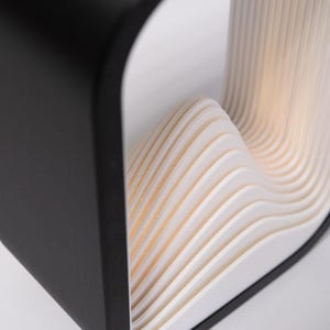 May include: A table lamp featuring a matte black outer frame with a square profile. The interior contains a series of stacked, white, wave-like layers that create a rhythmic, undulating pattern. The structure emits a soft, warm light from within.