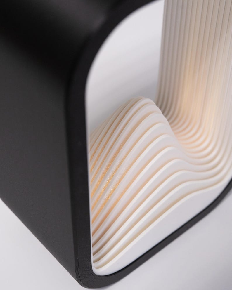 May include: A table lamp featuring a matte black outer frame with a square profile. The interior contains a series of stacked, white, wave-like layers that create a rhythmic, undulating pattern. The structure emits a soft, warm light from within.