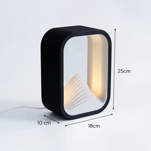 May include: A black rectangular table lamp with a white interior featuring a layered mountain-like structure. The lamp emits a warm light and measures 25 cm in height, 18 cm in width, and 10 cm in depth, as indicated by the visible dimension labels.