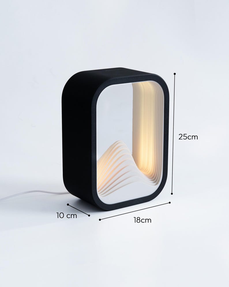 May include: A black rectangular table lamp with a white interior featuring a layered mountain-like structure. The lamp emits a warm light and measures 25 cm in height, 18 cm in width, and 10 cm in depth, as indicated by the visible dimension labels.