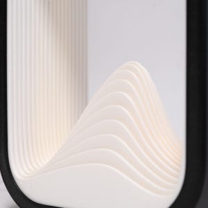 May include: A rectangular object with a black outer frame and a white interior featuring a series of layered, curved, wave-like ridges that rise to a central peak. The structure has a smooth, matte finish and a clean, geometric design.