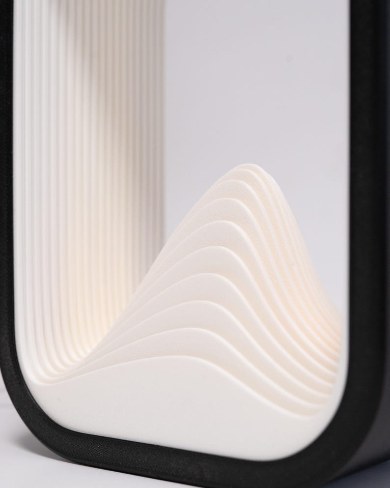 May include: A rectangular object with a black outer frame and a white interior featuring a series of layered, curved, wave-like ridges that rise to a central peak. The structure has a smooth, matte finish and a clean, geometric design.