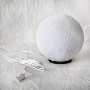 May include: A white, spherical lamp with a textured surface, resembling the moon. The lamp sits on a black base and has a white cord with a USB connector. The lamp is designed for decorative lighting.