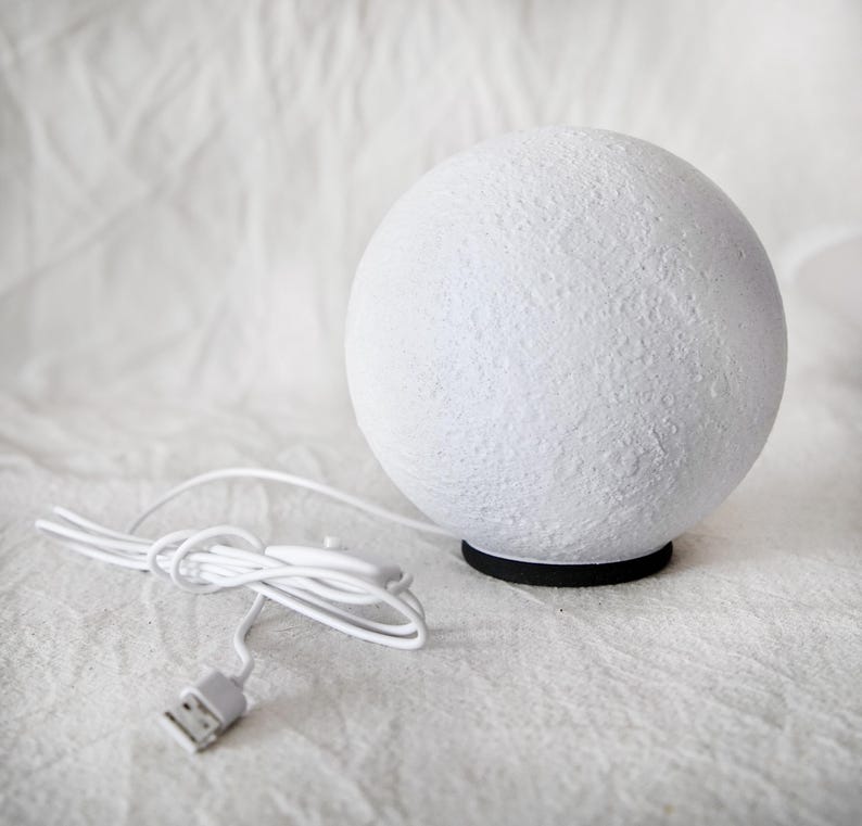 May include: A white, spherical lamp with a textured surface, resembling the moon. The lamp sits on a black base and has a white cord with a USB connector. The lamp is designed for decorative lighting.