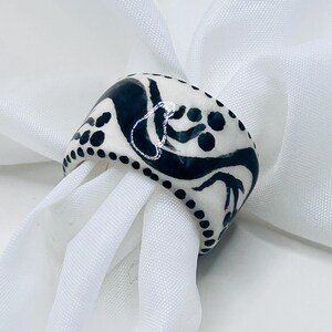 May include: A ceramic ring with a white base and black abstract designs. The ring features a glossy finish and is adorned with black dots along the edges. The ring is displayed on a white fabric.