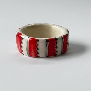 May include: A ceramic ring with a bold design. The ring features alternating vertical stripes of red and white, accented with silver and small black dots. The interior of the ring is a cream color.