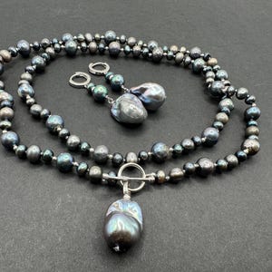 May include: A necklace and earring set featuring dark gray and iridescent pearls. The necklace has a toggle clasp and a large, teardrop-shaped pearl pendant. The earrings have a similar design with large, baroque pearls and silver hoops.