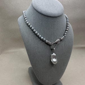 May include: A gray pearl necklace displayed on a gray bust. The necklace features a strand of small, round pearls and a larger, baroque pearl pendant. The clasp is embellished with small, sparkling stones. The necklace is a piece of jewelry.