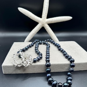 May include: A black pearl necklace with a silver clasp featuring a decorative design. The necklace is displayed on a gray surface with a white starfish in the background. The pearls have a lustrous sheen.