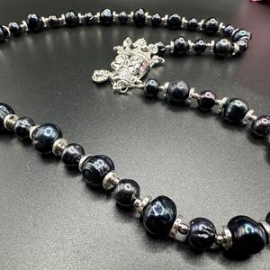 May include: A black pearl necklace with a silver clasp. The necklace features a variety of dark, lustrous pearls, interspersed with small silver beads. The clasp is an ornate silver design. The necklace is displayed on a dark surface.