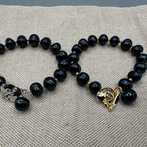 May include: Two black pearl bracelets with gold and silver clasps. The bracelets are on a beige fabric background.