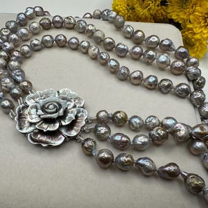 May include: A multi-strand pearl necklace with a large, silver-toned flower pendant. The pearls are a mix of gray and iridescent colors. The flower pendant has detailed petal designs. Yellow flowers are in the background.