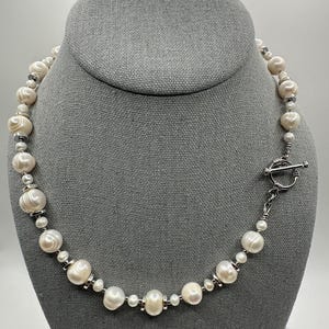 May include: A pearl necklace displayed on a gray bust. The necklace features large, off-white pearls interspersed with smaller, silver-toned beads and accents. It has a toggle clasp closure.