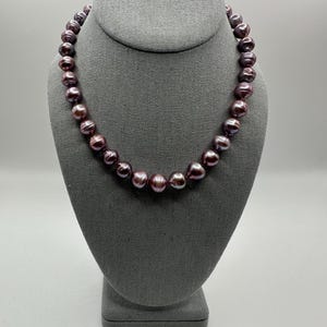 May include: A necklace of dark purple pearls displayed on a gray jewelry bust. The pearls have a lustrous sheen and are strung together to form a complete necklace. The jewelry bust is made of a soft, textured fabric.