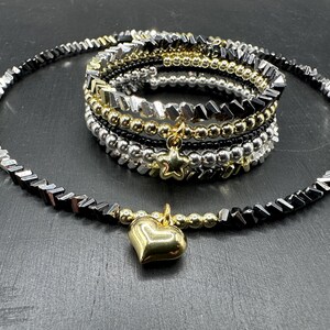 Könnte beinhalten: Eine schwarze und silberne Perlenkette mit einem goldenen Herz-Anhänger. Die Kette ist mit mehreren Perlensträngen geschichtet, darunter schwarze, silberne und goldene Perlen.