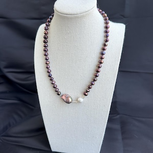 May include: A necklace featuring a strand of dark, iridescent pearls. The necklace has a large, irregularly shaped pearl and a smaller, round white pearl as a focal point. The pearls are strung on a silver-colored chain, displayed on a white mannequin.