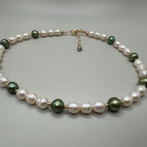 May include: A necklace featuring alternating white and dark green pearls. The necklace has a gold-colored clasp and connecting elements. The pearls are round and vary slightly in size, creating a classic and elegant design.