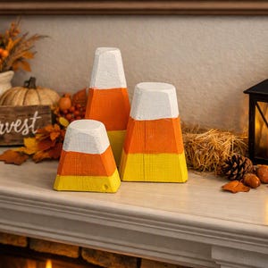 May include: Three decorative candy corn figures crafted from wood, each featuring horizontal bands of white, orange, and yellow paint. The figures are arranged in a cluster, showcasing a smooth, matte finish with visible wood grain texture.