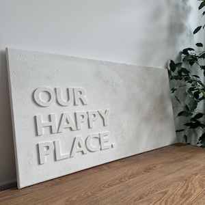 May include: A rectangular white wall art piece featuring the raised, embossed text "OUR HAPPY PLACE." The surface has a textured, matte finish with subtle variations in tone, creating a three-dimensional effect for the lettering against the panel.
