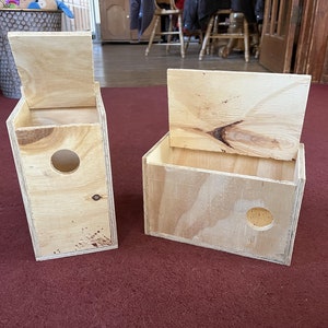 Puede incluir: Dos casas para pájaros de madera sin terminar con tapas con bisagras. Una es más alta y estrecha, la otra es más corta y ancha. Cada una tiene un agujero de entrada circular. Las casas para pájaros están hechas de madera contrachapada de color claro.