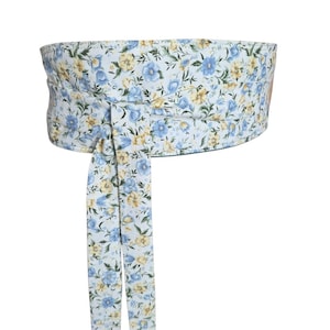 May include: A wide wrap belt featuring a white background with a repeating floral pattern in shades of light blue and yellow. The belt has a structured, cylindrical main section and two long, narrow fabric ties hanging down from the front center.
