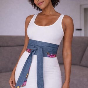 Puede incluir: Una mujer con un vestido blanco de tirantes acanalado y un cintur&oacute;n envolvente de mezclilla. El cintur&oacute;n tiene un forro con estampado floral. El vestido es entallado. El cintur&oacute;n est&aacute; atado con un lazo en la cintura.