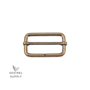 May include: A gold-toned metal buckle with a rectangular shape and a single bar across the center. The buckle is designed to be used for straps or belts.