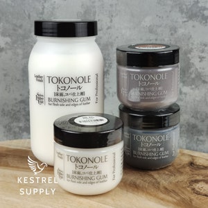 Può includere: Un assortimento di prodotti di gomma per lucidatura Tokonole per la lavorazione della pelle. L'immagine mostra una grande bottiglia bianca e tre barattoli più piccoli in vari colori, tutti con coperchi neri ed etichette di prodotto. Il testo sulle etichette include "TOKONOLE" e "BURNISHING GUM".