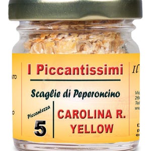 Op de afbeelding: Een kleine glazen pot met een gouden deksel gevuld met gele pepervlokken. De pot heeft een geel etiket met de tekst "I Piccantissimi Scaglie di Peperoncino CAROLINA R. YELLOW" en een pittigheidsclassificatie van 5.