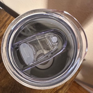 May include: A clear plastic tumbler lid with a sliding closure. The lid is transparent, revealing the interior of the tumbler. The lid is designed to fit snugly on a tumbler, providing a spill-resistant seal. The tumbler is sitting on a wooden surface.