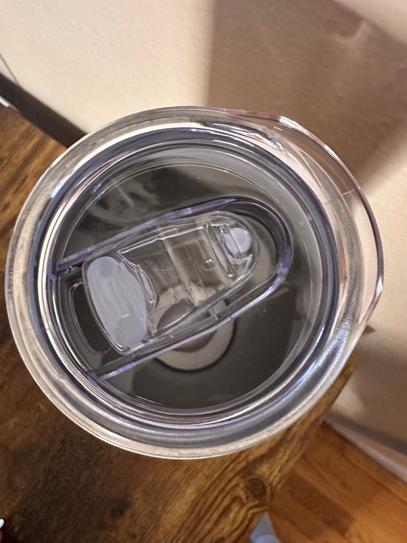 May include: A clear plastic tumbler lid with a sliding closure. The lid is transparent, revealing the interior of the tumbler. The lid is designed to fit snugly on a tumbler, providing a spill-resistant seal. The tumbler is sitting on a wooden surface.