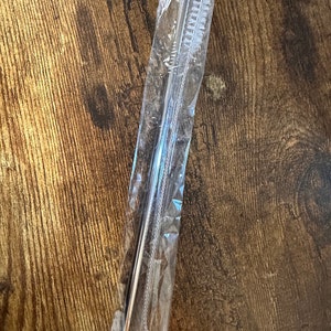 May include: A set of stainless steel straws and a cleaning brush, packaged in clear plastic. The straws are straight and cylindrical. The brush has white bristles. The items are on a wooden surface.