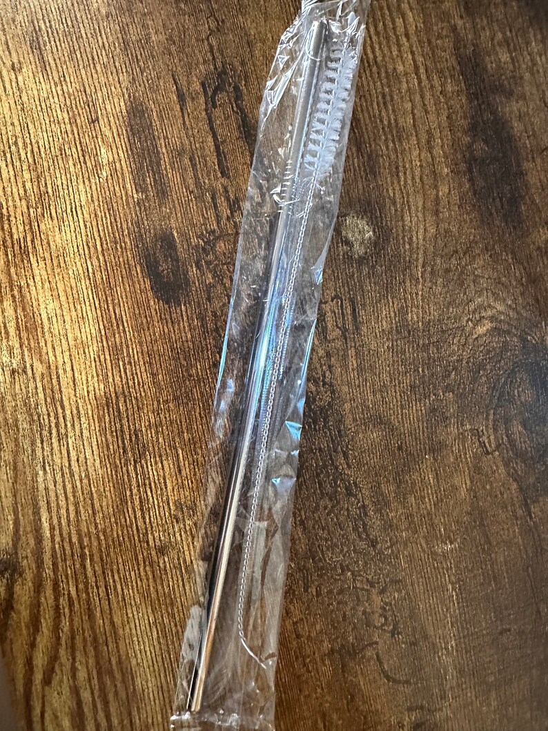 May include: A set of stainless steel straws and a cleaning brush, packaged in clear plastic. The straws are straight and cylindrical. The brush has white bristles. The items are on a wooden surface.