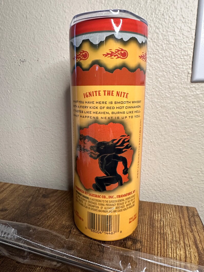 May include: A cylindrical tumbler with a clear lid, featuring a fiery design and the text "IGNITE THE NITE." The tumbler has a yellow background with orange flames and a silhouette of a figure. The text describes the drink as smooth whisky with cinnamon.