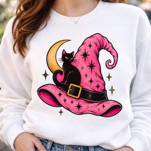 Könnte beinhalten: Ein weißes Sweatshirt mit Rundhalsausschnitt und einem Grafikdruck eines rosa Hexenhuts mit schwarzem Gürtel und Schnalle. Eine schwarze Katze sitzt auf dem Hut vor einem gelben Halbmond. Das Design enthält verstreute schwarze und goldene Sterne.