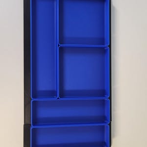 May include: A rectangular drawer organizer featuring a black outer frame and five distinct blue compartments of varying sizes. The layout includes one long vertical section, two square sections, and two horizontal sections for storage.