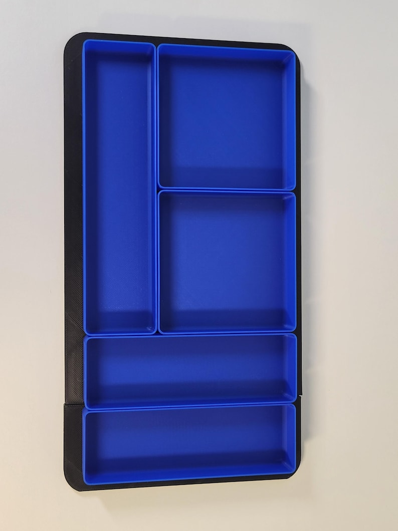 May include: A rectangular drawer organizer featuring a black outer frame and five distinct blue compartments of varying sizes. The layout includes one long vertical section, two square sections, and two horizontal sections for storage.