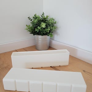 May include: Two rectangular, off-white planters are on a wooden floor. One has square indentations. The other is behind it. A small potted plant with green leaves is in the background, in a silver metal container.