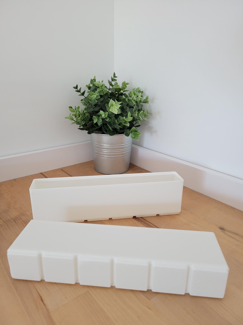 May include: Two rectangular, off-white planters are on a wooden floor. One has square indentations. The other is behind it. A small potted plant with green leaves is in the background, in a silver metal container.