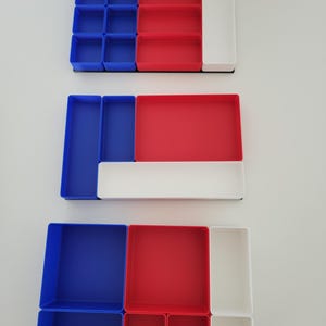 May include: Three rectangular drawer organizer sets featuring modular compartments in solid blue, red, and white plastic. Each set contains various sizes of open-top bins arranged in different configurations to provide segmented storage for small items.