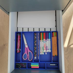 May include: A gray desk drawer containing blue plastic organizers filled with office supplies. The contents include pink-handled scissors, pens, patterned pencils, colorful markers, thin colored pens, paper clips, sticky notes, and a battery.