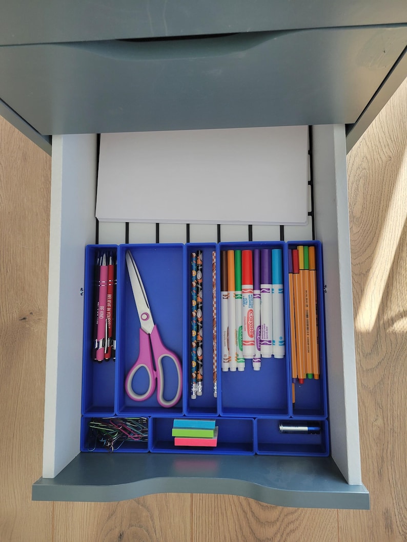May include: A gray desk drawer containing blue plastic organizers filled with office supplies. The contents include pink-handled scissors, pens, patterned pencils, colorful markers, thin colored pens, paper clips, sticky notes, and a battery.
