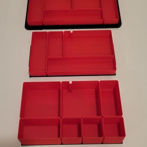 May include: Three rectangular red plastic drawer organizers with multiple compartments of varying sizes. Each organizer features a black base and interior dividers, providing structured sections for sorting small items within a drawer or storage space.