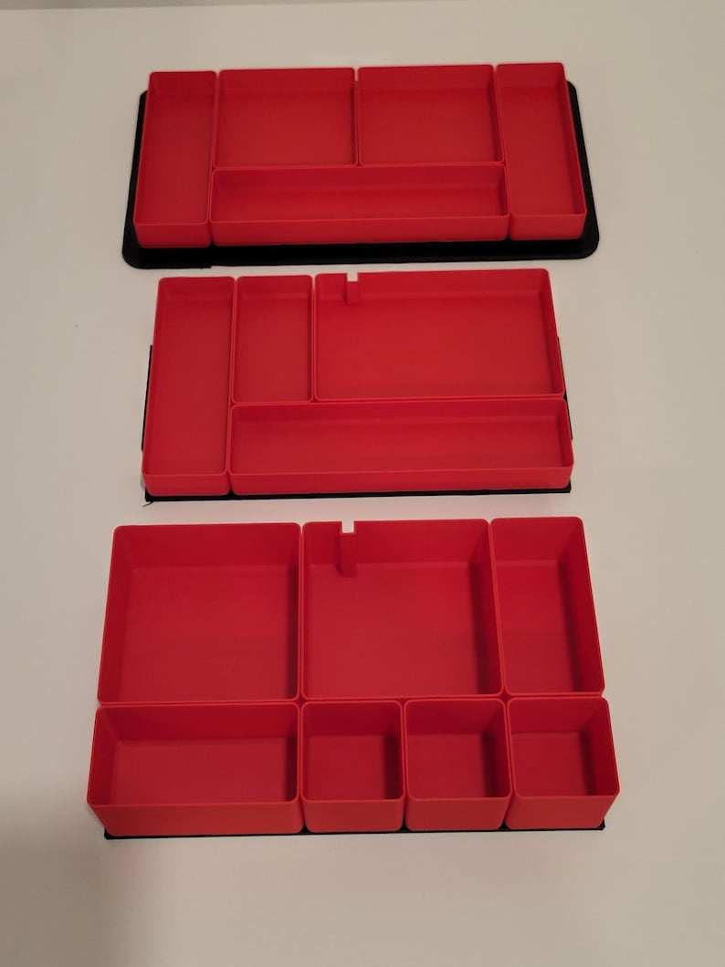 May include: Three rectangular red plastic drawer organizers with multiple compartments of varying sizes. Each organizer features a black base and interior dividers, providing structured sections for sorting small items within a drawer or storage space.