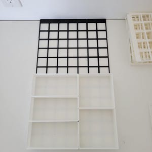 May include: A modular drawer organizer system featuring a black grid base plate and several white rectangular bins of varying sizes. The white bins are arranged to fit into the grid, creating multiple compartments for storage and organization.