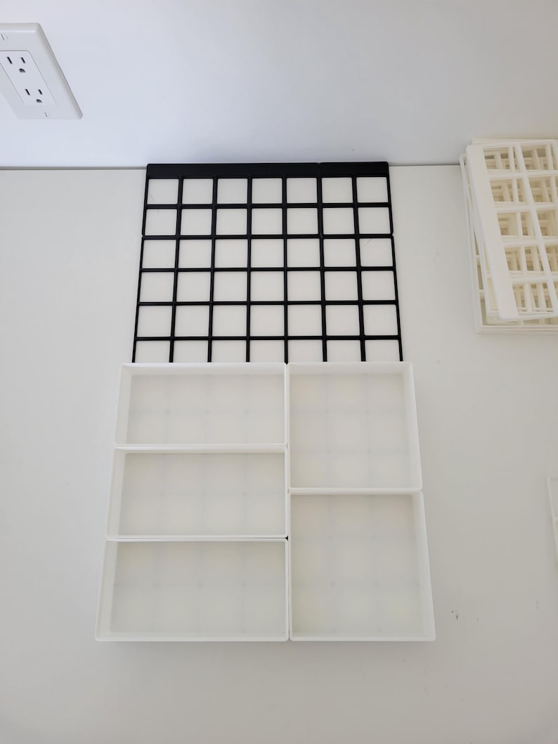 May include: A modular drawer organizer system featuring a black grid base plate and several white rectangular bins of varying sizes. The white bins are arranged to fit into the grid, creating multiple compartments for storage and organization.